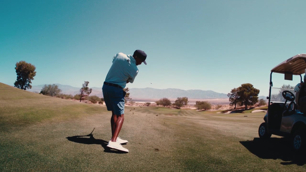 Golfista di colore esegue un colpo netto con il ferro dal fairway in ultra slow motion mentre la telecamera si inclina verso il basso, con un campo da golf tra montagne desertiche sullo sfondo, 4k 800 fps in estate