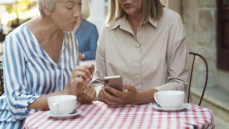 Zwei-Gut-Aussehende-ältere-Damen-Sitzen-Am-Tisch-Auf-Der-Caféterrasse-Und-Sehen-Sich-Fotos-Auf-Dem-Smartphone-An