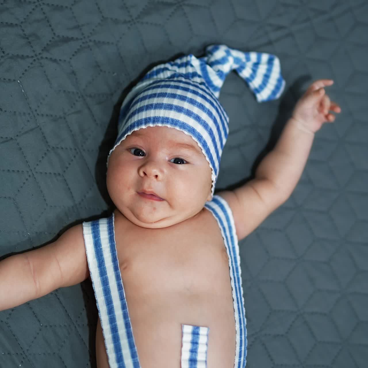 Sweet boy wearing striped suit lies on the bed with his hands sideways. Funny costume on a kid at the grey background