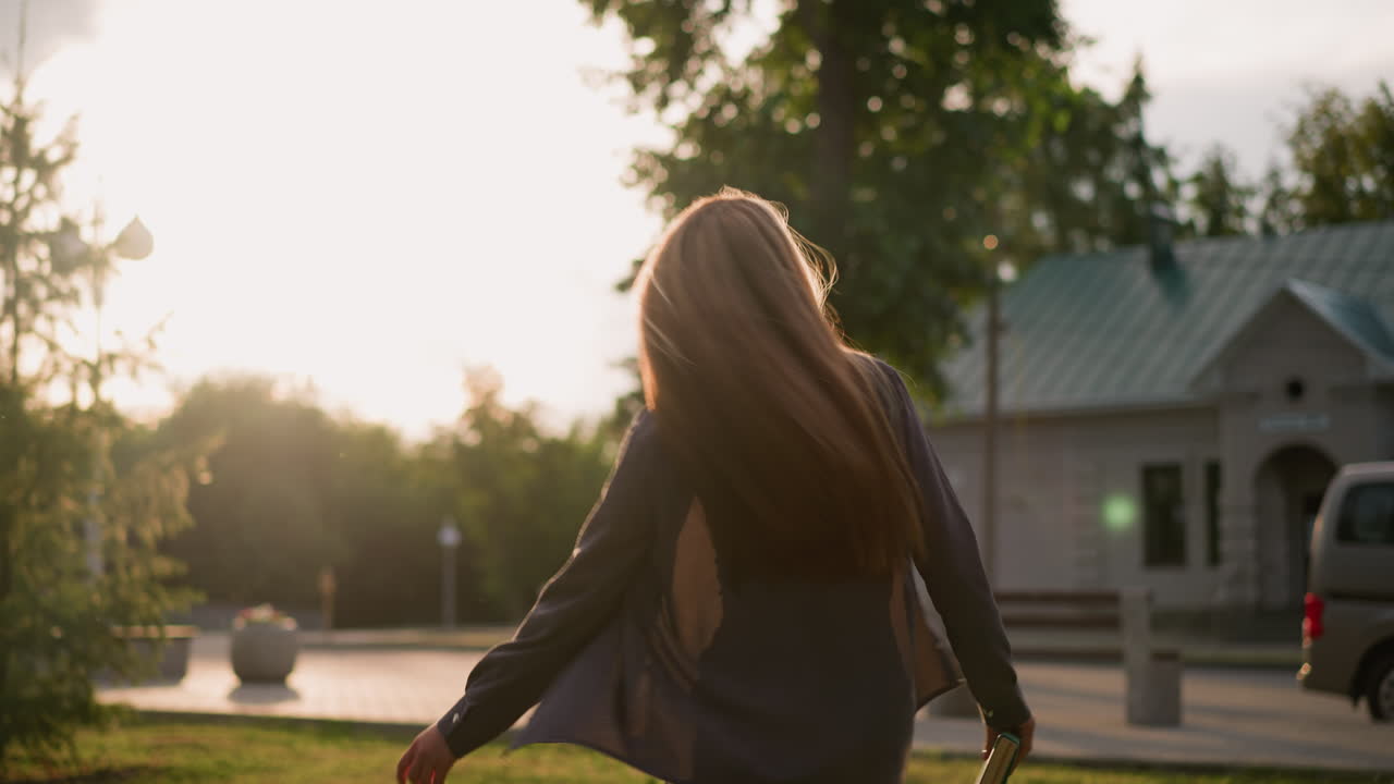 한 여인이 책 한 권을 들고 잔디에 있는 야외에서 즐겁게 춤을 추고 있는데, 빛은 그녀를 둘러싸고 반이는 분위기를 조성하고, 배경에는 초록색과 나무와 건물이 있습니다.