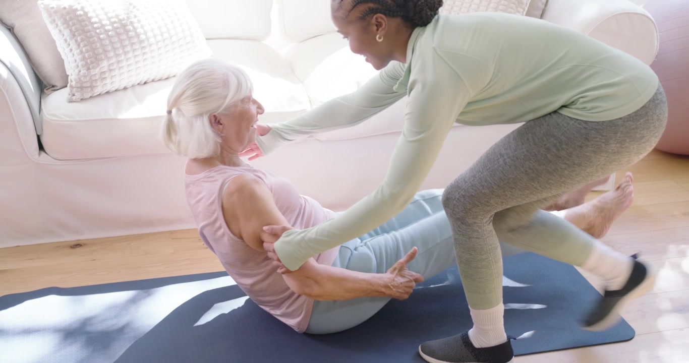 Senior woman lying on mat in living room, trainer bending knee guiding sit up for mobility