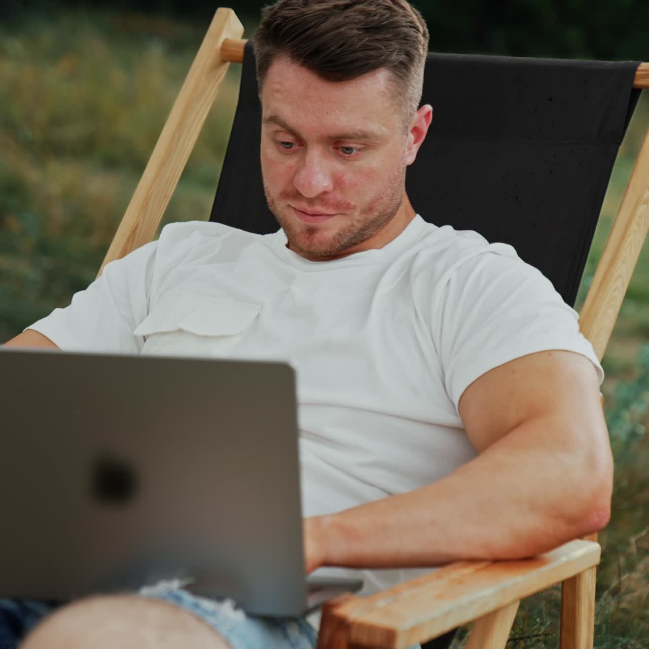 Freelancing Caucasian man works outdoors. Man sits in a folding chair focused on the laptop screen