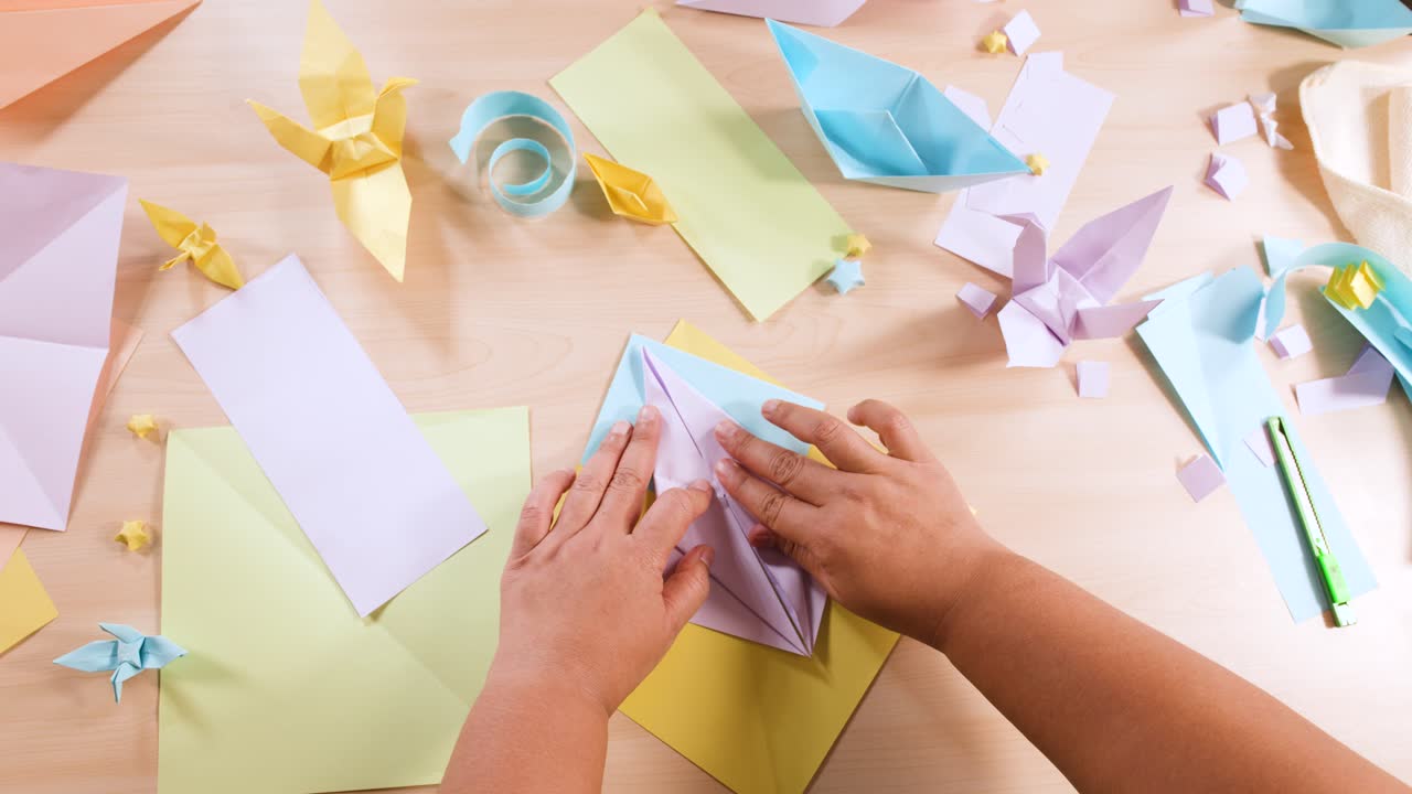 Visuale dall'alto di mani che piegano con cura carta viola su una scrivania piena di creazioni origami