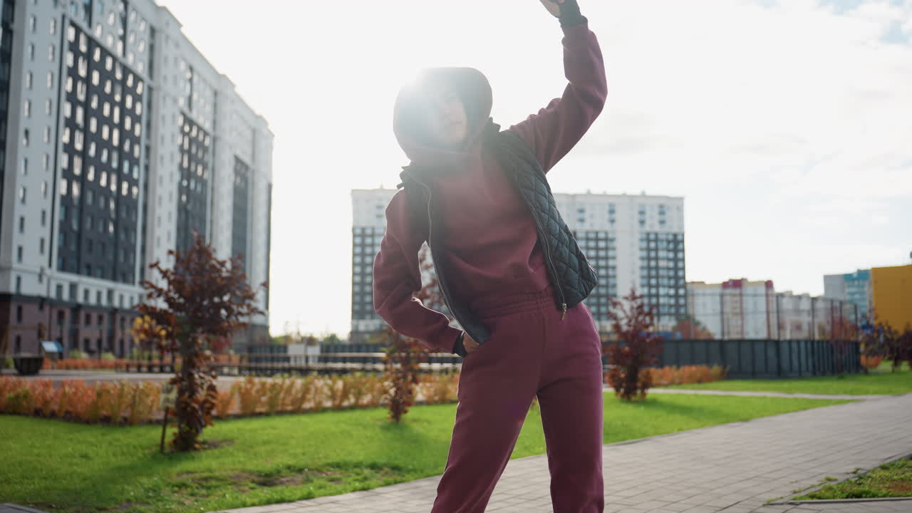 Energetic young woman in hoodie and black jacket stretches side muscles outdoors under bright sun on city plaza surrounded by high rise apartments and autumn foliage during urban fitness routine