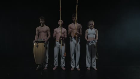 Group of a professional dancers are practicing capoeira in darkness against a spotlight on a black background of studio. Afro-Brazilian martial art that combine elements of dance.