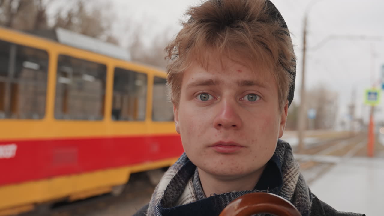 Weißer Kreativer Fotograf blickt auf ankommende Straßenbahn, rote und gelbe Straßenbahn fährt auf nassen Gleisen vorbei, entschlossener Blick, Schal und Mütze, Regenschirmgriff sichtbar, Nahaufnahme Porträt, urbane Bewegung.