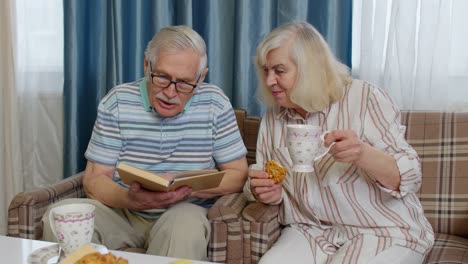 smiling senior couple grandfather, grandmother resting on sofa drinking coffee, reading book at home