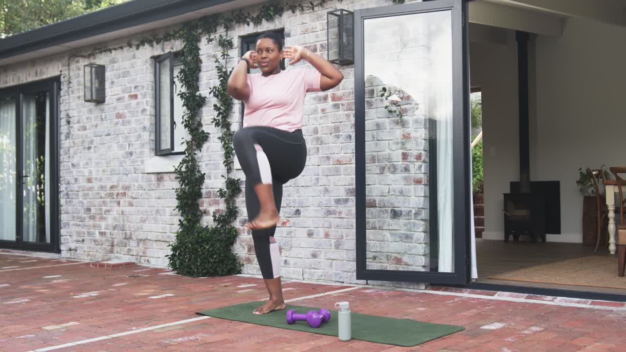 African American woman lifting arms doing high knees on red brick patio with yoga mat, dumbbells