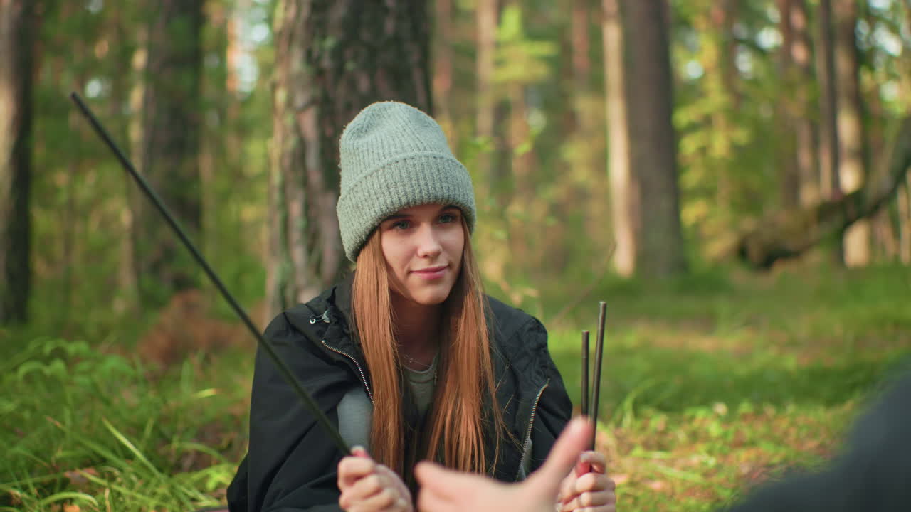 Woman holding tent poles while smiling softly and playfully delaying handover to her fiance in forest, enjoying relaxed camping moment surrounded by trees and soft sunlight filtering through woods