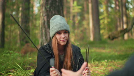woman holding tent poles while smiling softly and playfully delaying handover to her fiance in forest, enjoying relaxed camping moment surrounded by trees and soft sunlight filtering through woods