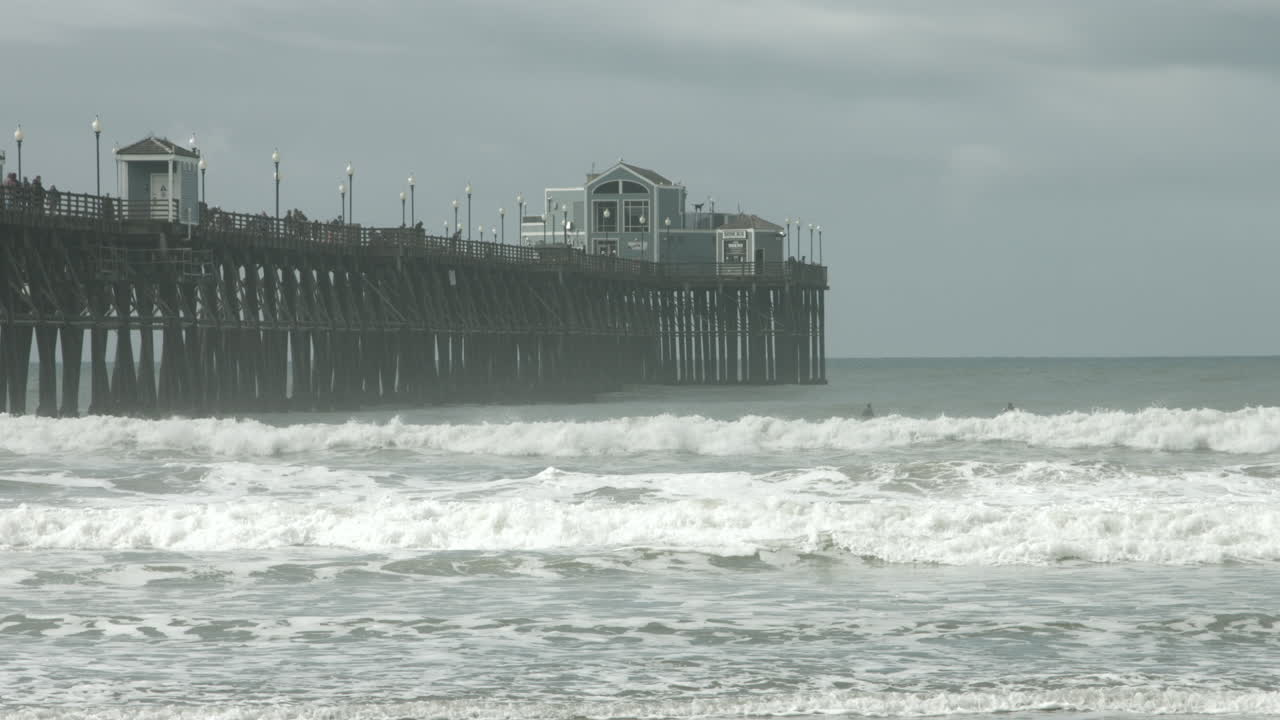 UHD footage of Oceanside Pier with dramatic waves crashing against the pilings beneath cloudy skies, showcasing the striking contrast between serene weather and the ocean's powerful force.