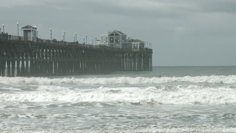 uhd footage of oceanside pier with dramatic waves crashing against the pilings beneath cloudy skies, showcasing the striking contrast between serene weather and the ocean's powerful force.