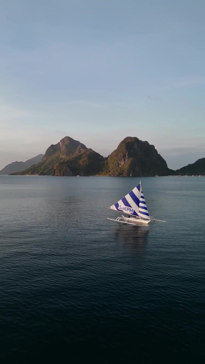Pemandangan udara drone yang tenang dari perahu cadik (paraw) tradisional berlayar bergaris meluncur di perairan laut yang tenang dengan pegunungan pulau tropis yang dramatis di latar belakang, El Nido, Filipina