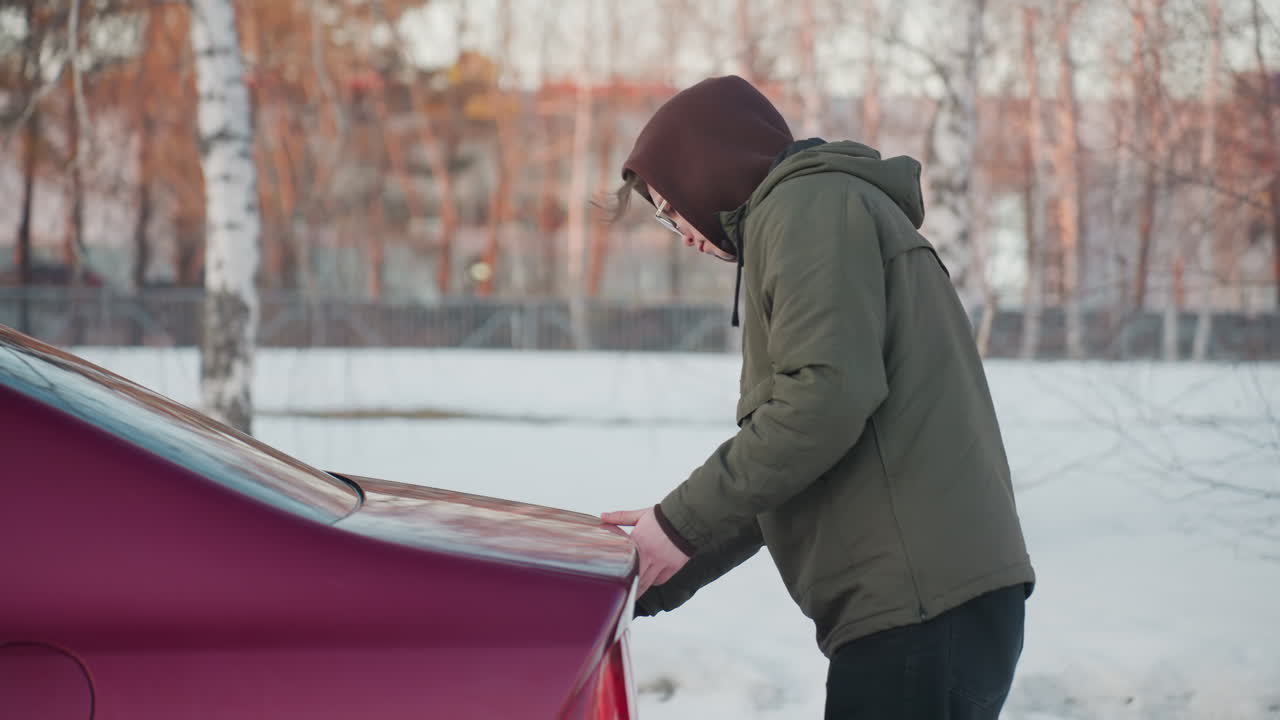 Teenager in hooded jacket opens red car boot on snowy ground with winter trees in background, side view showing effort to lift trunk, overcast lighting and soft natural tones in cold environment