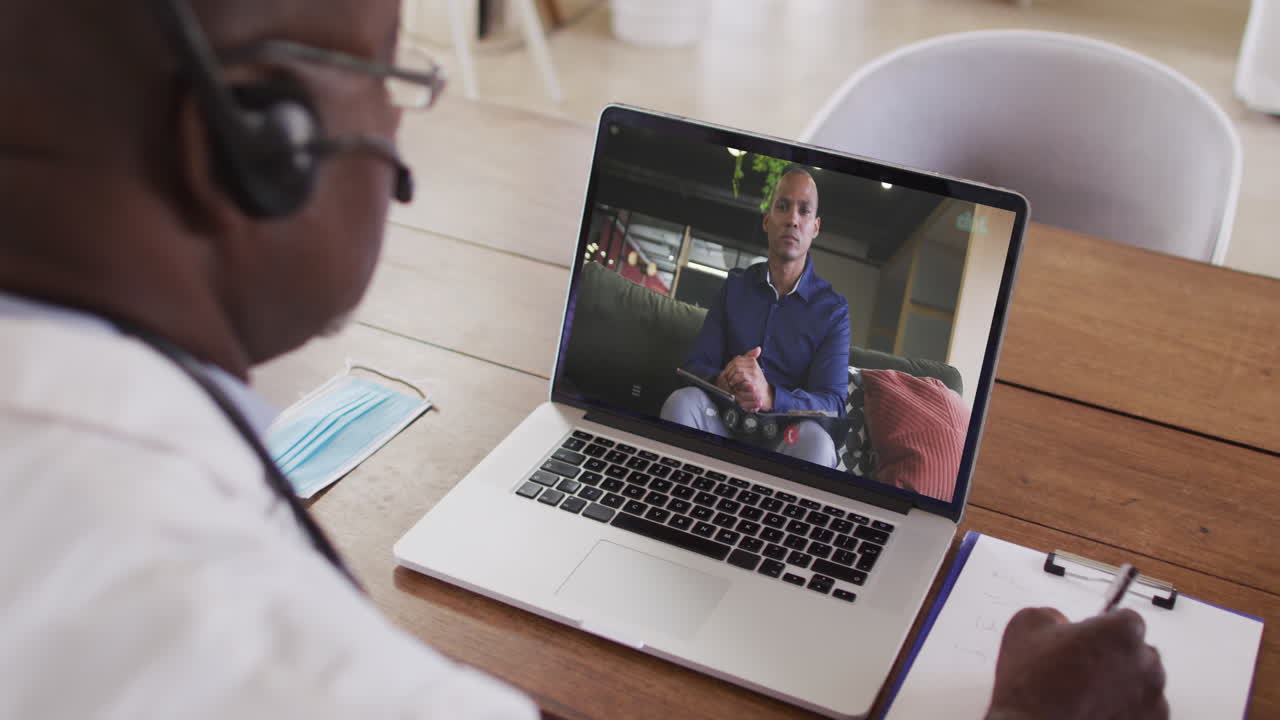 Afroamerikansk mandlig læge med headset på, der tager noter under et videoopkald på sin bærbare computer