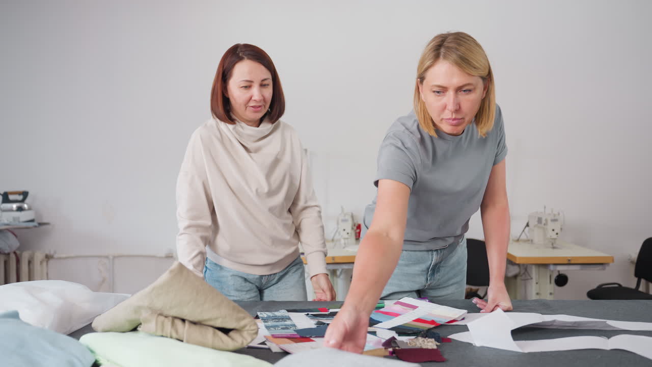 Seamstress unfolding plain material while reviewing colorful fabric samples on table, preparing for fashion project in creative workspace filled with sewing tools, and garment patterns