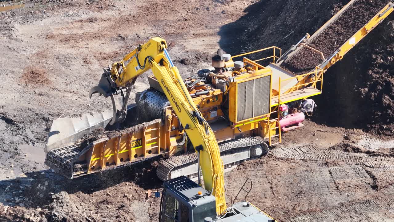 Drone captures an excavator loading soil into a processing machine, creating mulch piles under bright daylight