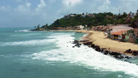 dolly out tilt up aerial drone shot of the tropical famous baia formosa beach town in the state of rio grande do norte, brazil with fishing boats, coastal homes, small waves, and surfers