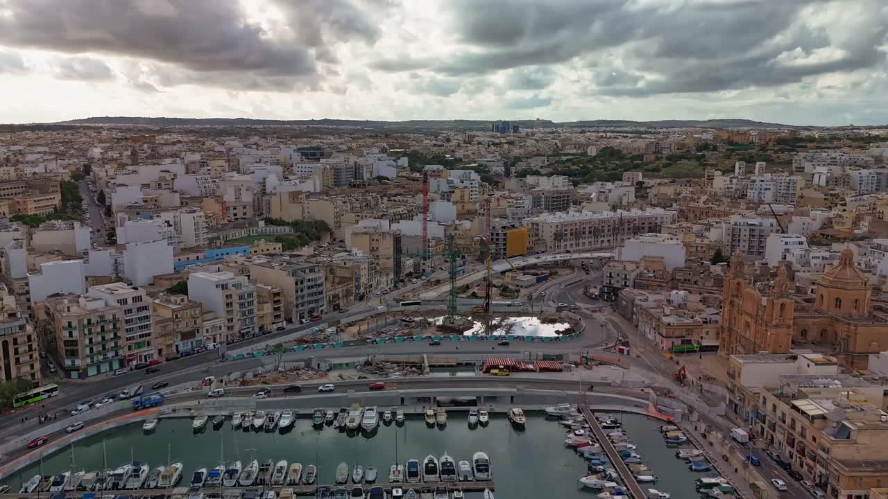 Aerial timelapse over Msida, Malta showing ongoing flyover construction beside the marina and St. Joseph Church, with urban traffic and dramatic clouds moving across the skyline