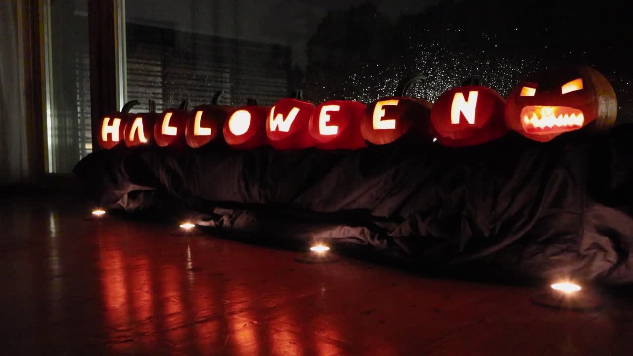 Carved out pumpkins in front of a window during night time. Pumpkins spell out Halloween with candles glowing and burning inside of them. Candles in front that reflect on yellow and orange light below