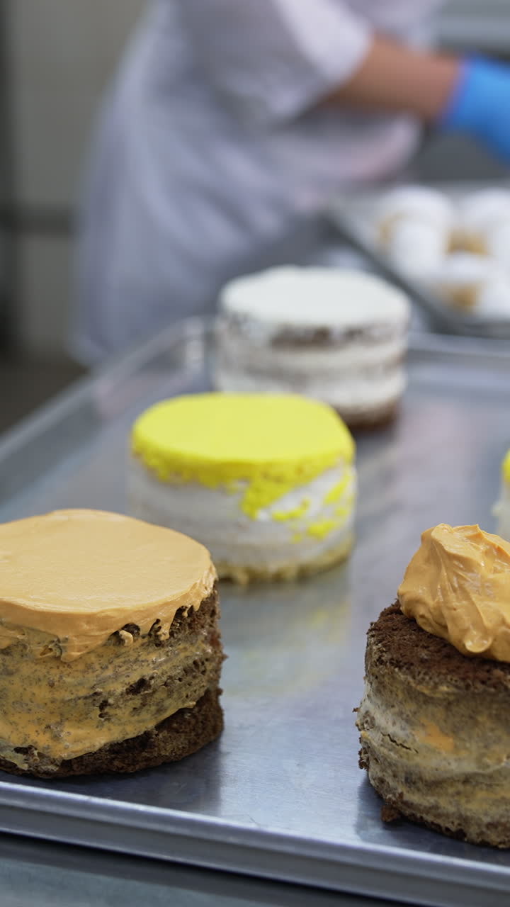 Metal spatula is used to spread cream over the cake surface. Varied cakes are placed on metal tray for decoration. Close up. Vertical video