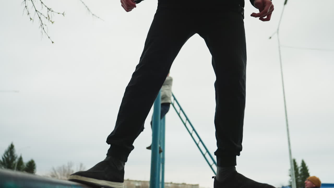 Close-up leg view of someone walking carefully on an iron rail, with a blurred view of another person in gray clothing climbing a blue pole in the background