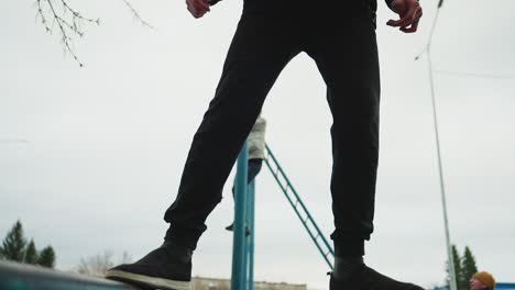 close-up leg view of someone walking carefully on an iron rail, with a blurred view of another person in gray clothing climbing a blue pole in the background