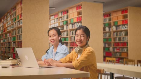 asian woman students with headphones typing on a laptop and writing into the notebook smiling to camera while sitting on a table studying in the library