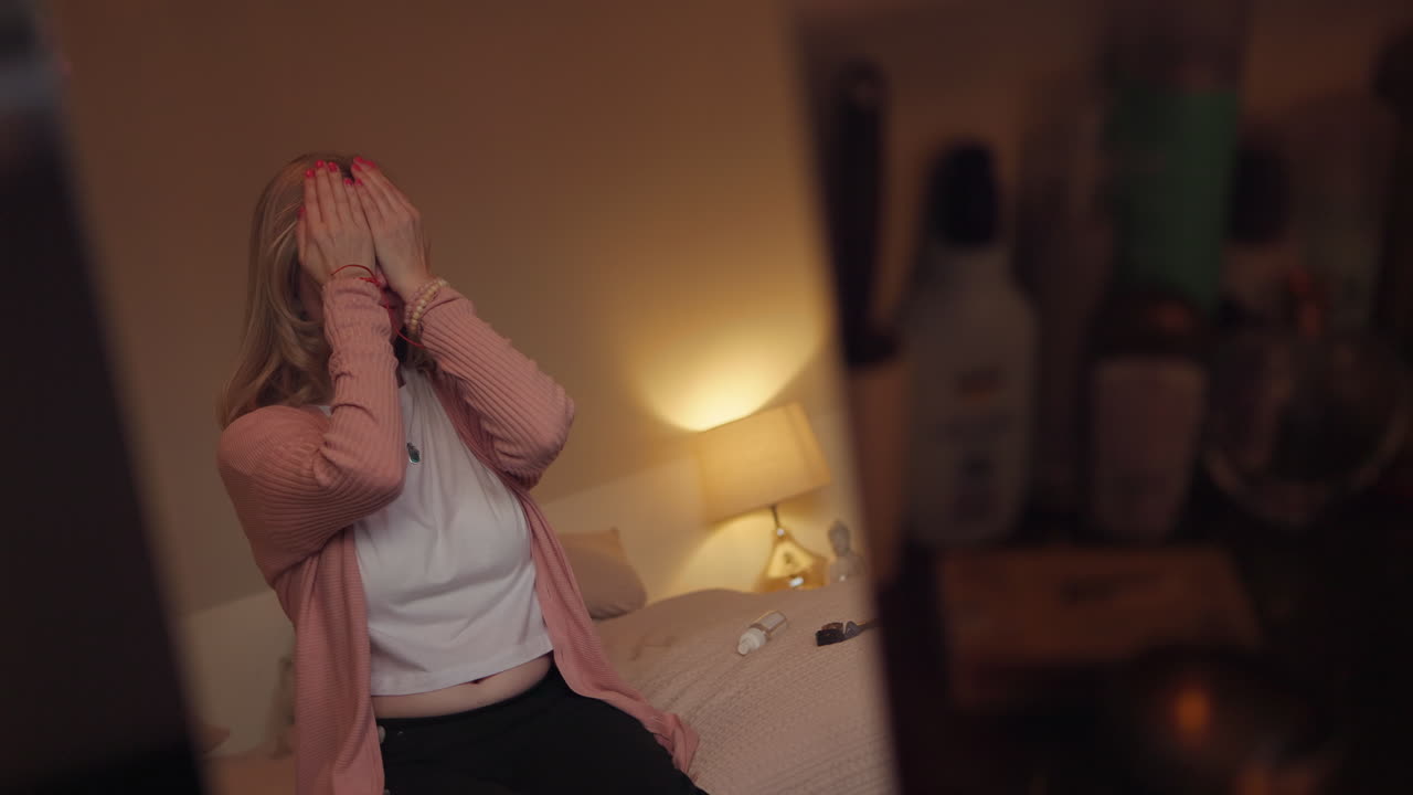 Woman in pink cardigan covering her face in front of a mirror in bedroom