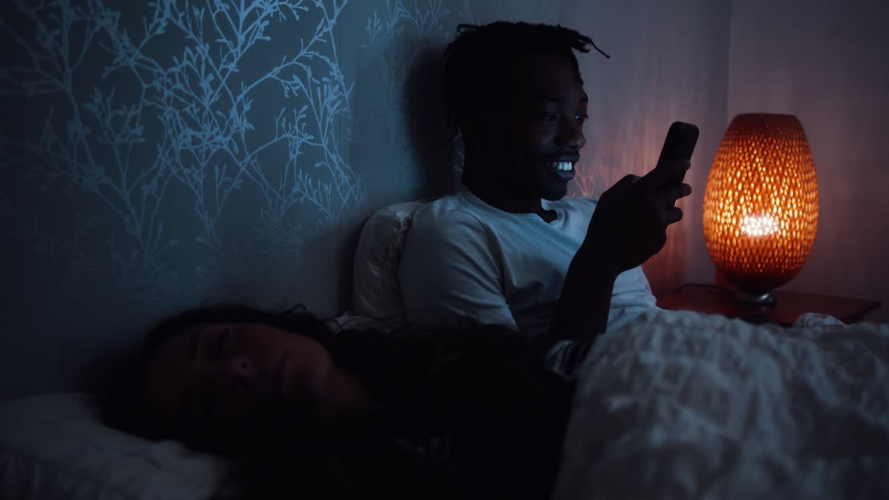 Giovane uomo sorride e guarda lo smartphone mentre è sdraiato a letto di notte.