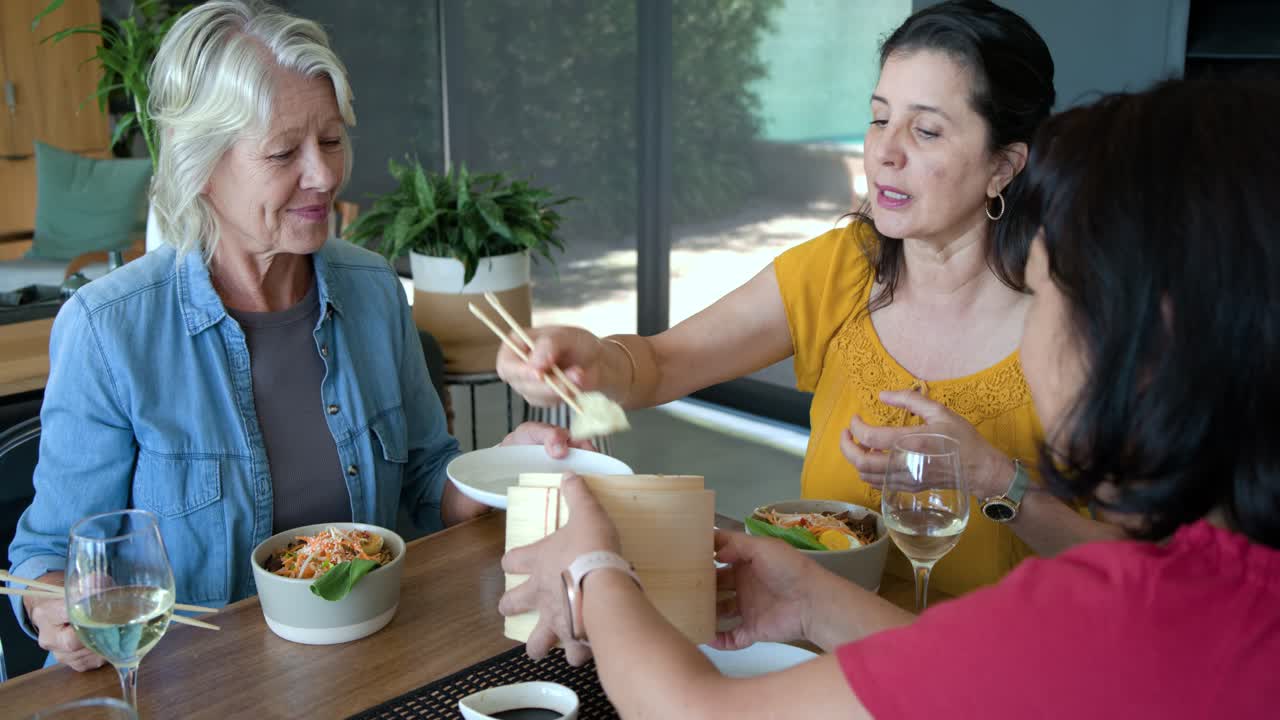 Mujeres diversas compartiendo una comida en casa, una anciana y una mujer de rojo sirviendo dim sum de una vaporera de bambú.