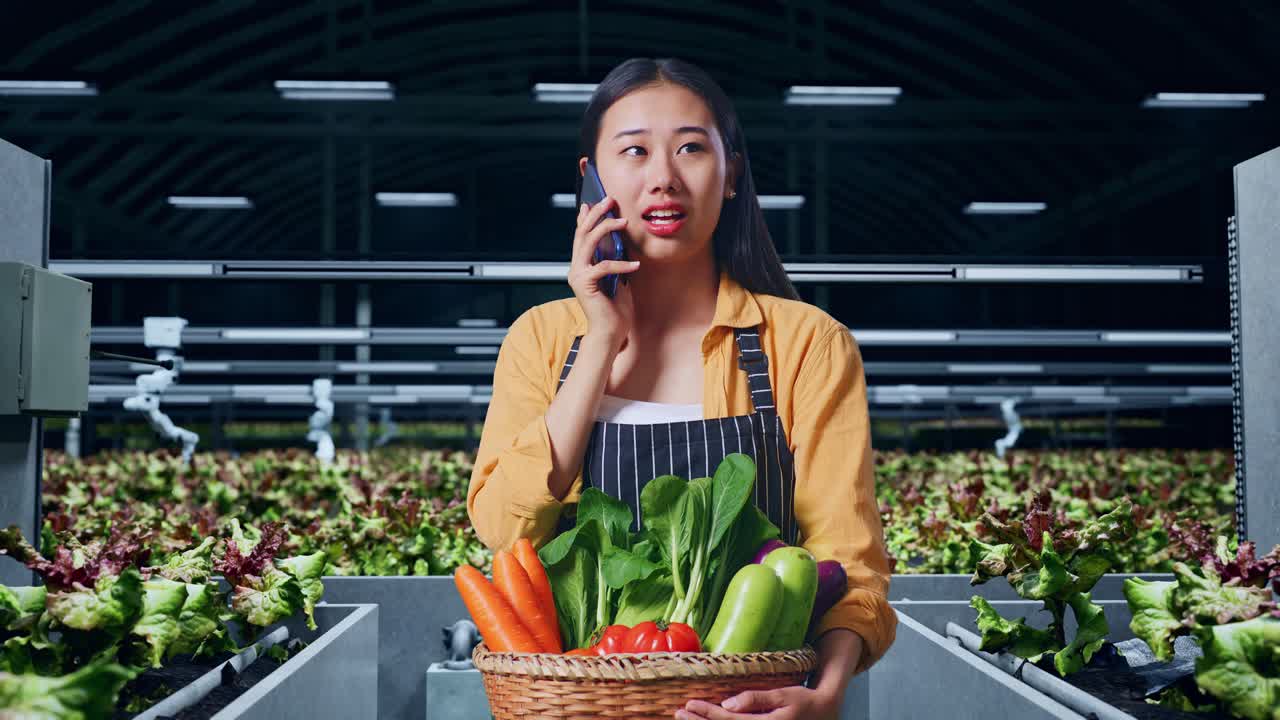 웃는 아시아 여성 농부 가 채소 바구니 를 들고 스마트 농장 에서 휴대 전화 로 이야기 하고 있다