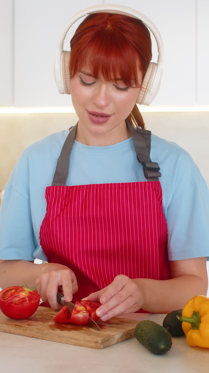 Young woman with headphones plays anti stress music cuts tomatoes for salad focused kitchen routine