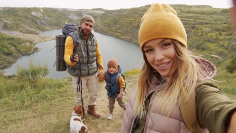 pov shot of happy young woman holding camera and taking selfie or filming herself and her cheerful husband, 5 year old son and cute jack russell terrier dog while hiking near quarry lake