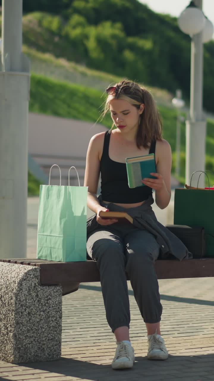 두 쪽 모두에 쇼핑 가방이 있는 야외 벤치에 앉아있는 여인이 민트 녹색 가방에서 책을 가져와 갈색 책을 같은 가방으로 돌려줍니다.