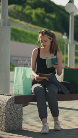 dama sentada en un banco al aire libre con bolsas de compras a ambos lados, recupera un libro de la bolsa verde menta y devuelve el libro marrón de nuevo en la misma bolsa, el fondo incluye postes de lámpara