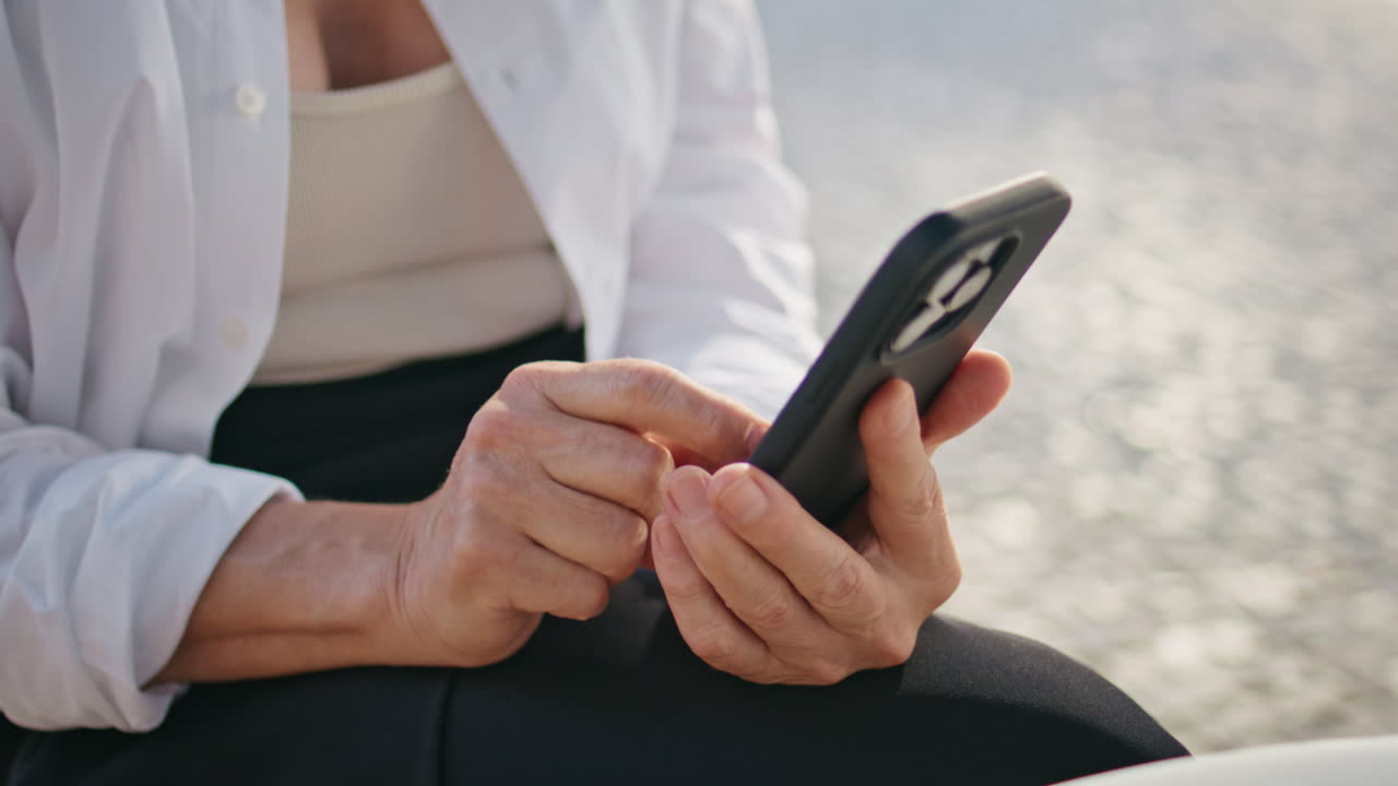 성숙한 손이 드폰 화면을 입력하고 길거리에서 가까이 앉아 있습니다. 나이 든 여자가 메시지를 보내고 있습니다.