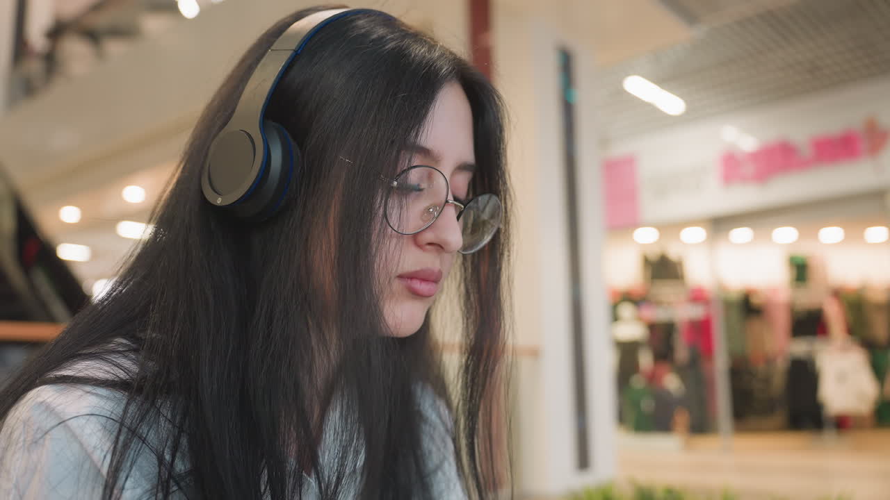 Close up of calm reflective woman with long dark hair and glasses seated in indoor mall space wearing headset, eyes closed while enjoying music, with blurred clothing store background