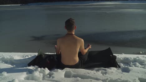 man with no shirt sitting on black yoga mat in snow beside frozen lake meditating