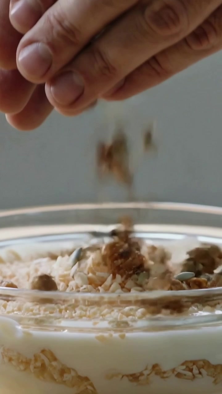 Video verticale: Mani adulte che si aprono e versano granola in una ciotola di vetro a casa, guarnendo lo yogurt.