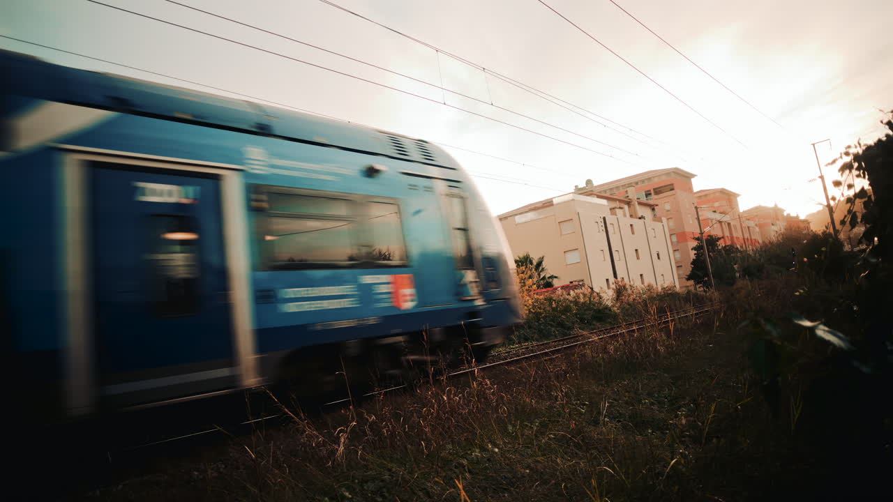 A blue commuter train speeds past a residential building, creating strong motion blur against the still architecture