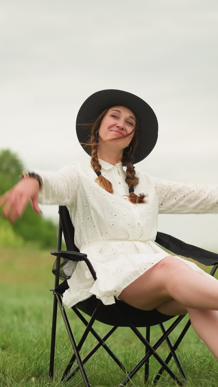 una mujer con un vestido blanco y un sombrero negro se sienta en una silla en un campo cubierto de hierba, agitando alegremente las manos en el aire y posando con una sonrisa brillante escena al aire libre captura un momento animado y despreocupado