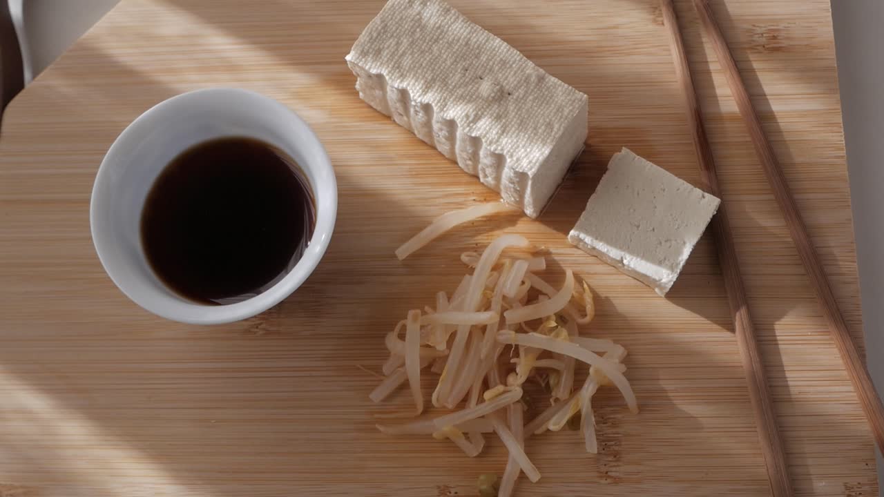 Tofu being sliced on wooden board, with bean sprouts, chopsticks and soy sauce in sunlight