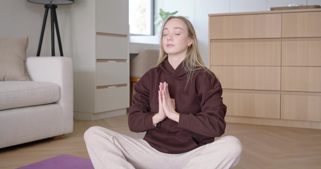 Donna a gambe incrociate su un tappetino da yoga viola in salotto, inspira e alza i palmi per la meditazione.