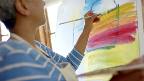 Focused-senior-biracial-woman-wearing-apron-and-painting-on-big-canvas-at-home,-slow-motion