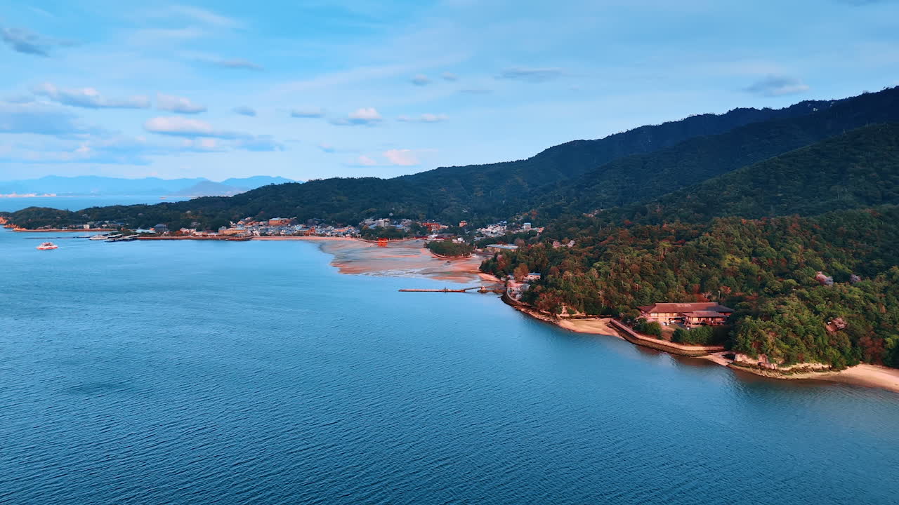 Flying above the tranquil blue waterscape approaching the famous red gates of Itsukushima Shrine at low tide season. Amazing scenic view of the mountains wooded with trees at the shore of Inland Sea of Japan.