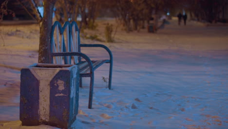 empty bench surrounded by snow under soft lighting with blurred figures walking in the distant background, creating a peaceful winter evening setting with serene, muted colors and shadows