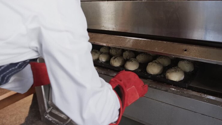 African american male baker working in bakery kitchen, putting rolls into oven in slow motion
