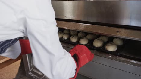 panadero afroamericano que trabaja en la cocina de la panadería, poniendo rollos en el horno en cámara lenta