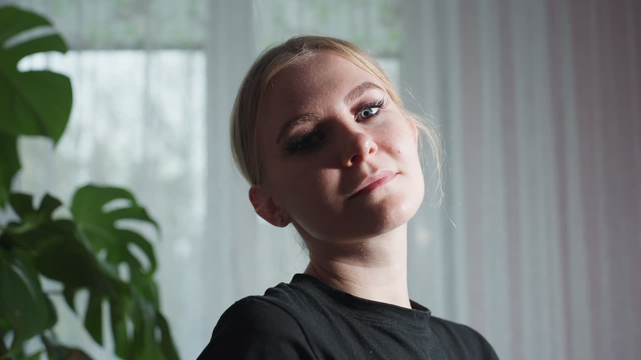 Close up of massage therapist in black shirt gazing at camera with serious expression, standing beside large leafy indoor plant, with soft daylight filtering through sheer curtains in background